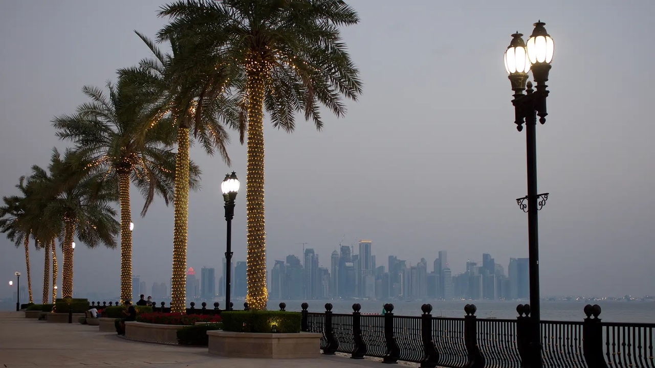 Doha Corniche promenade at twilight featuring illuminated palm trees, street lamps, and a distant city skyline.