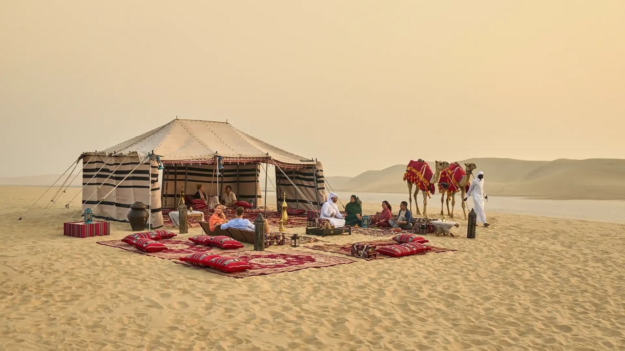 Guests relaxing at a traditional Bedouin camp with red rugs, cushions and camels on the golden sand dunes during a desert safari in Doha, Qatar