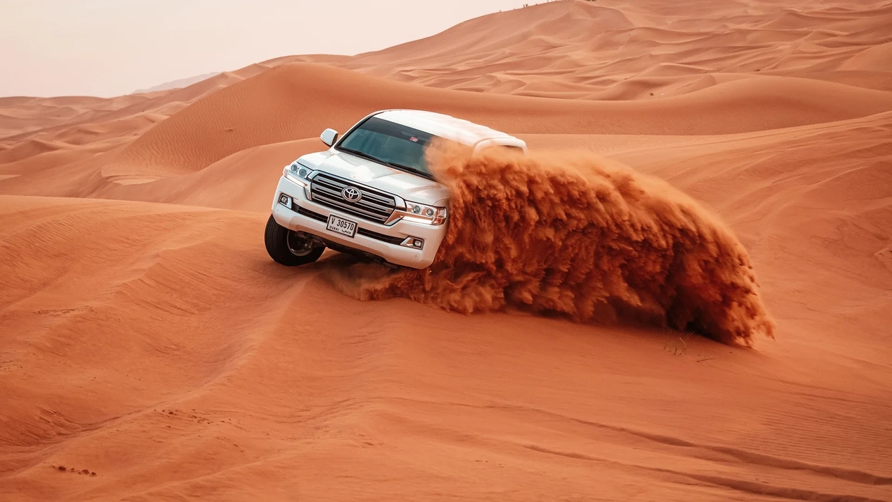 A white Toyota Land Cruiser dune bashing across red sand dunes with a dramatic sand spray during a desert safari in Doha, Qatar