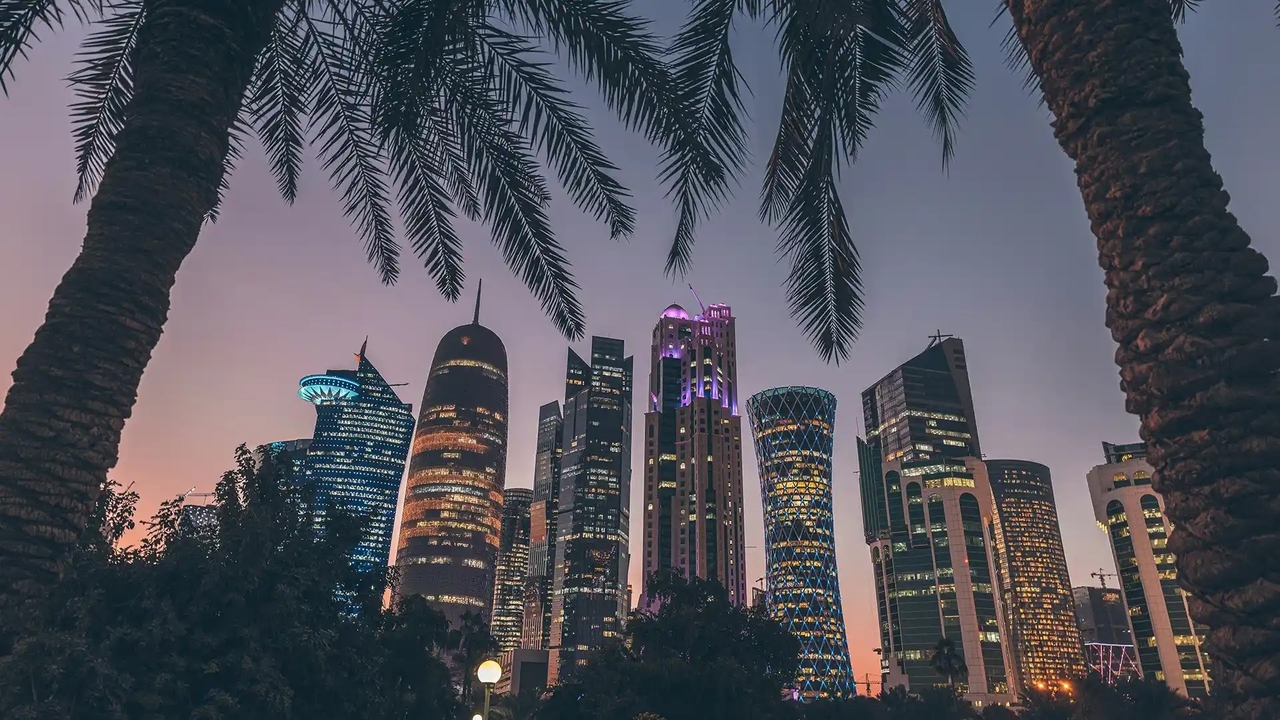 Modern skyscrapers of the Doha skyline lit up at dusk, framed by palm trees.