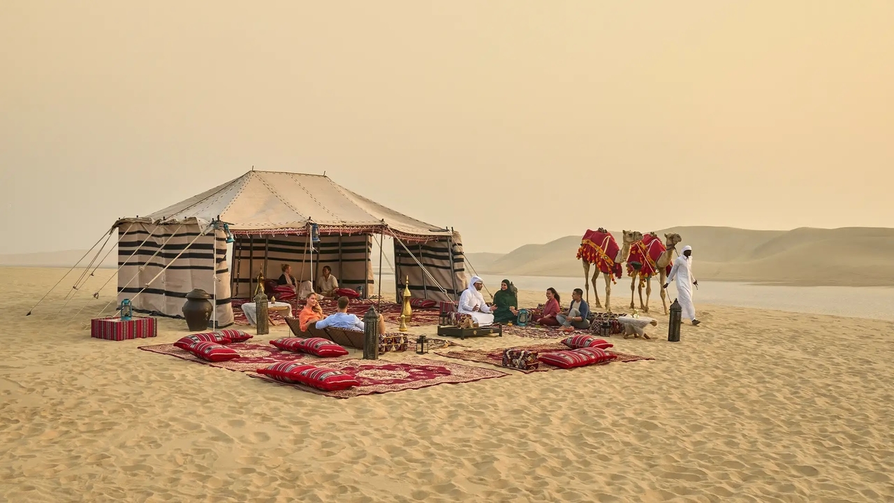 Guests relaxing at a traditional Bedouin camp with red rugs, cushions, and camels on the golden sand dunes during a desert safari in Doha, Qatar