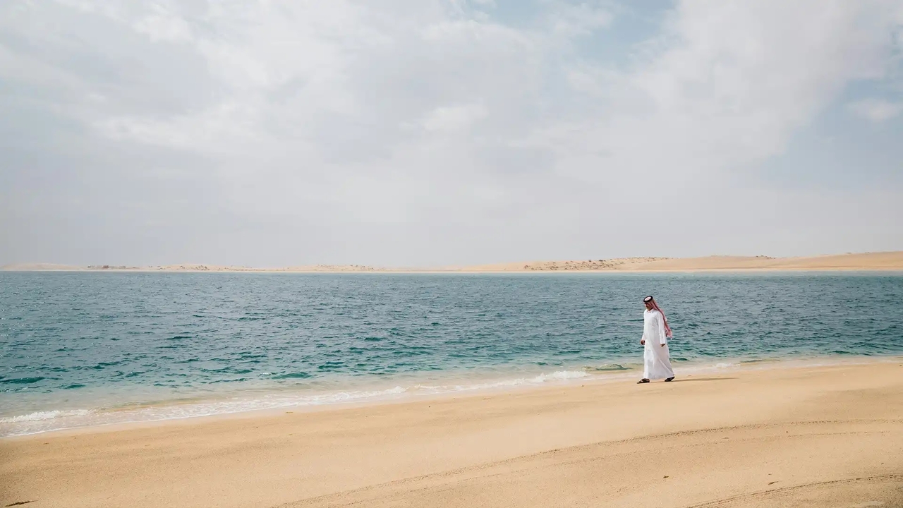 A man in traditional thobe walking along a quiet sandy beach next to turquoise Qatari waters.