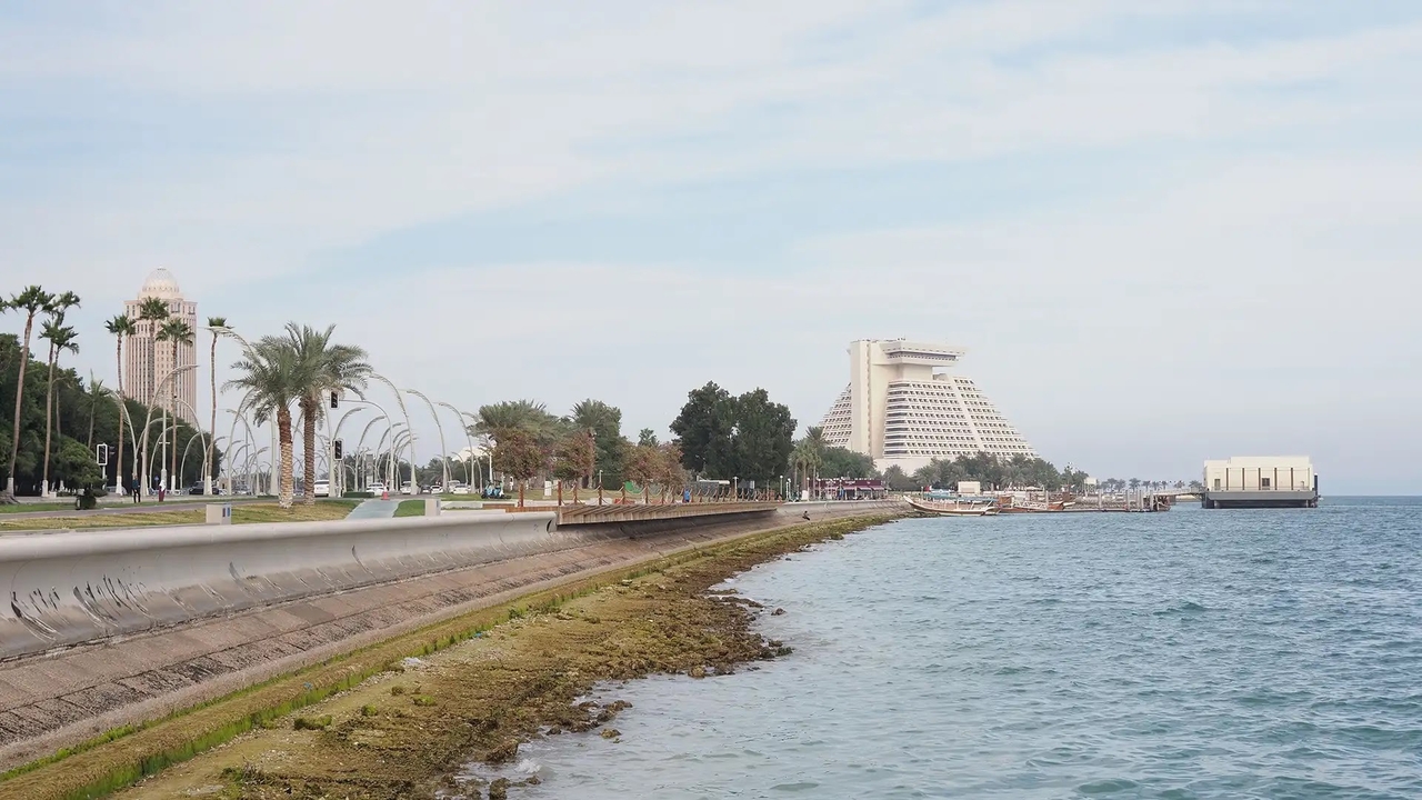 View of the Doha Corniche promenade, featuring palm trees, a sea wall, and the pyramid-shaped Sheraton hotel in the background.