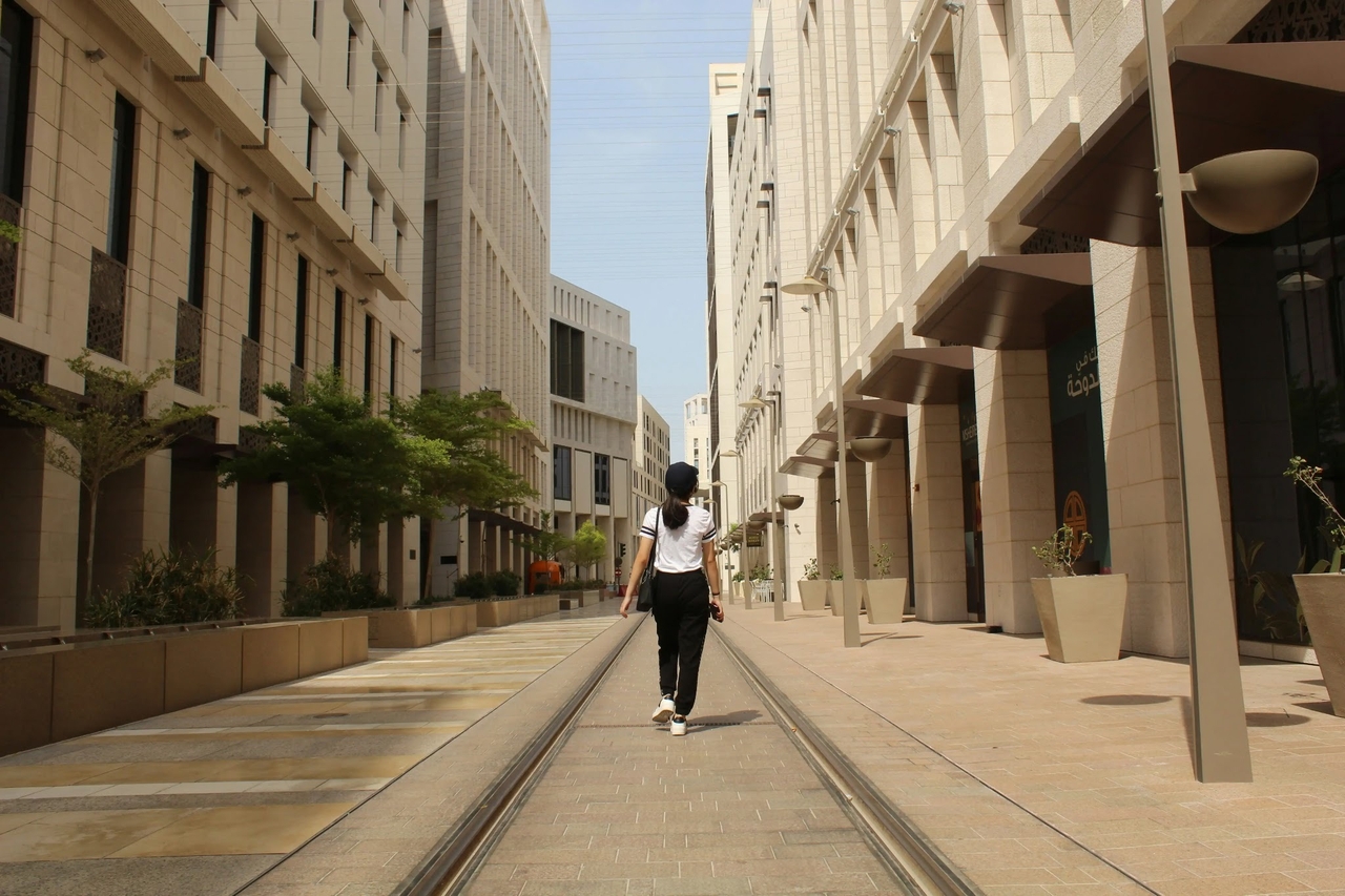 A person from behind walking along tram tracks in Msheireb Downtown Doha, flanked by modern stone buildings.