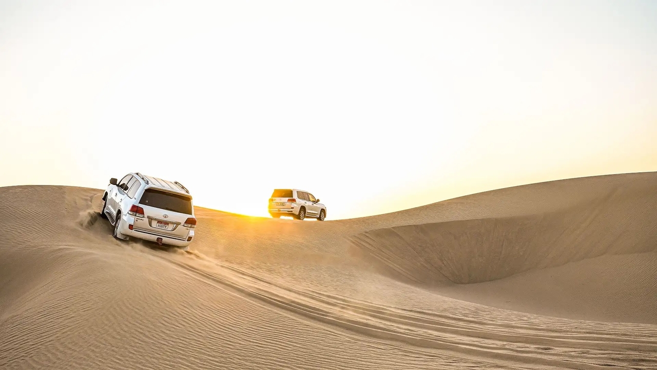 Two white Toyota Land Cruisers dune bashing across golden dunes during sunset on a desert safari in Doha, Qata